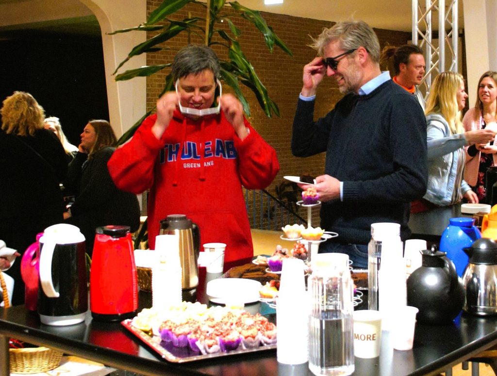 To publikummer prøver sanseudfordrende briller ved kaffebordet under Autonome Autister, mens andre deltagere står omkring dem.